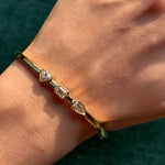 The Trio Bezel Set Mixed Shape Diamond Bangle in Yellow Gold shines on the wrist with heart, rectangular, and teardrop-shaped diamonds set side by side.