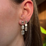 Close-up of a woman wearing Bezel Set Emerald Cut Diamond Drop Earrings in yellow gold, with each diamond elegantly bezel set and natural light highlighting the sparkling jewelry against a soft background.