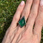 A hand with natural nails wears a Collet Set Kite Shaped Emerald Ring in black rhodium and yellow gold, with green grass in the background.