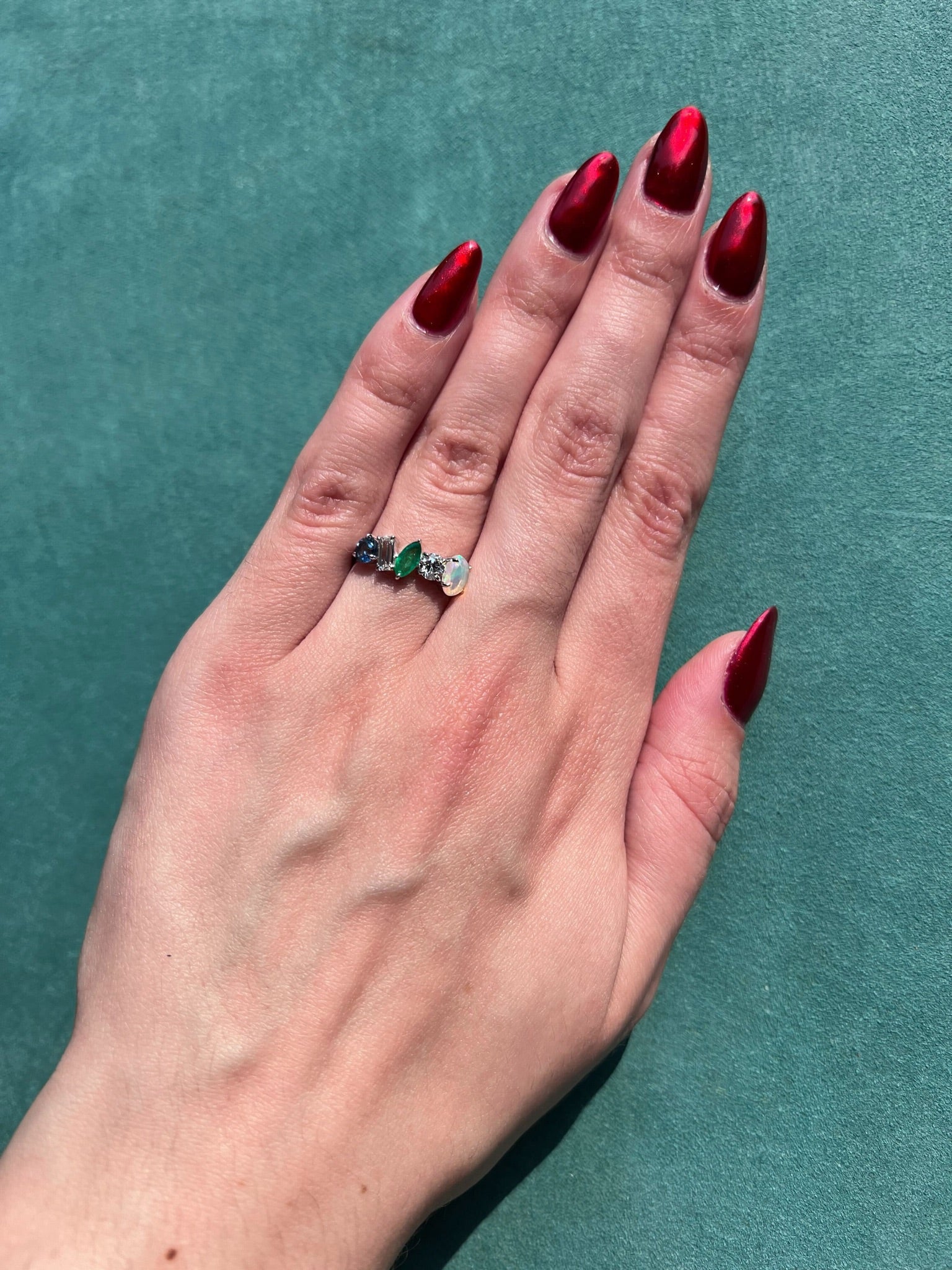 A hand with long, red metallic nails rests on a green surface, showcasing the Mixed Shape Opal, Emerald, Diamond, and Sapphire Half-Eternity Band in 18K white gold.