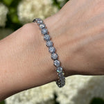 A close-up of a wrist wearing the Large Hexagon Bezel Set Diamond Tennis Bracelet, featuring hexagon-shaped settings with sparkling diamonds. The blurred background highlights light-colored flowers.