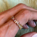 A manicured hand wears the Multicolor Eternity Band in yellow gold, featuring tapered baguette sapphires and round diamonds, set against a light green textured background.