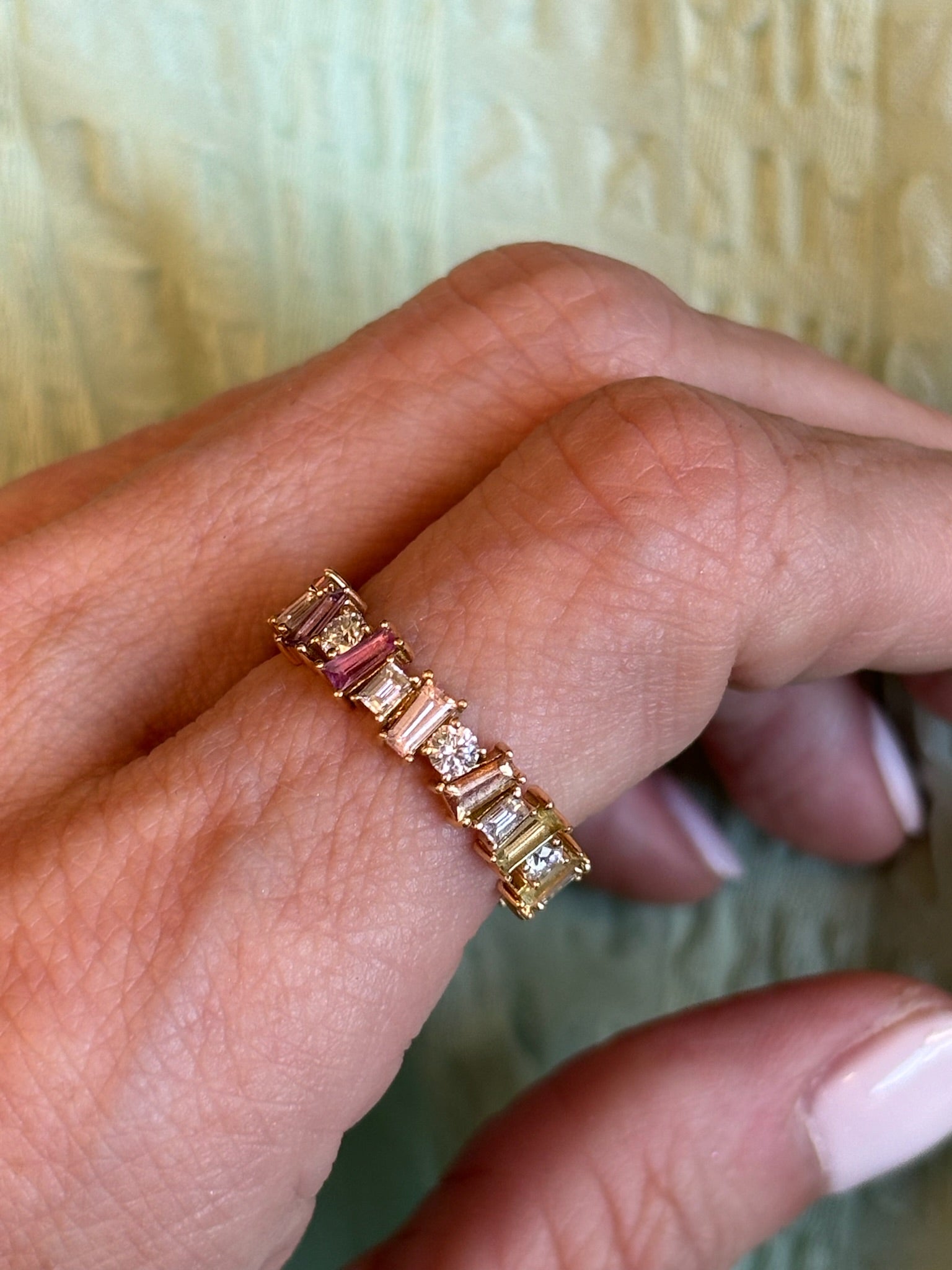 A manicured hand wears the Multicolor Eternity Band in yellow gold, featuring tapered baguette sapphires and round diamonds, set against a light green textured background.