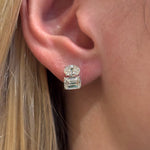 Close-up of an ear wearing the East-West Set Oval and Emerald Cut Diamond Studs in White Gold, featuring a round diamond above and a larger emerald-cut diamond below. The person's blonde hair is tucked behind the ear.