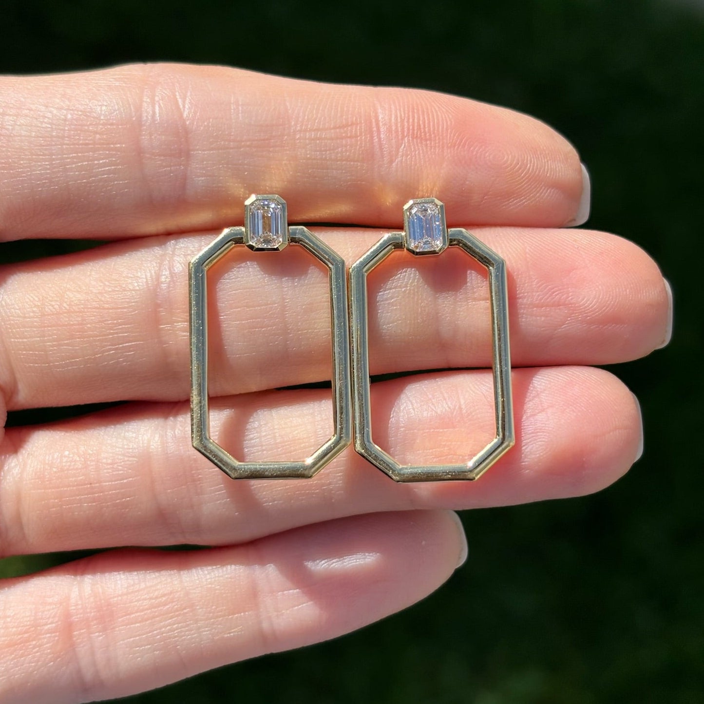 A hand holds Bezel Set Emerald Cut Diamond Earrings with an emerald motif hoop in yellow gold, featuring octagonal open frames and rectangular diamonds, against a blurred dark green background.
