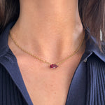 A person wears the East-West Pear Ruby Solitaire Pendant in Yellow Gold, its petite pear-shaped ruby resting above the collar of a dark textured shirt.