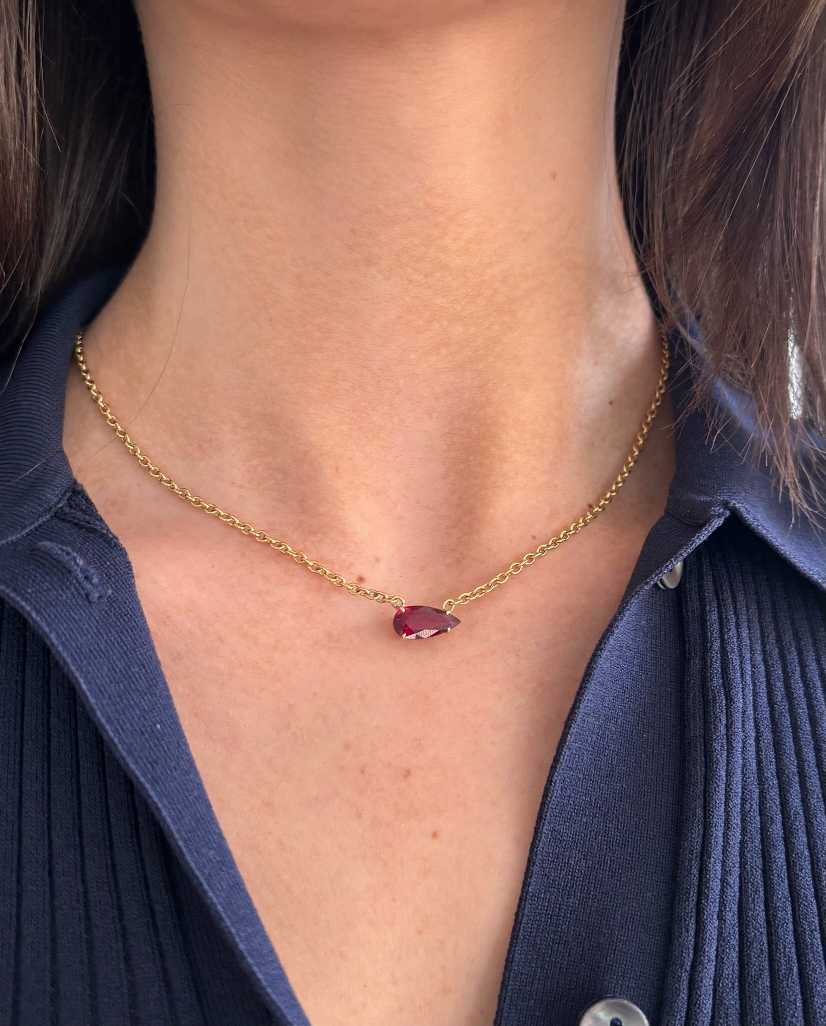A person wears the East-West Pear Ruby Solitaire Pendant in Yellow Gold, its petite pear-shaped ruby resting above the collar of a dark textured shirt.