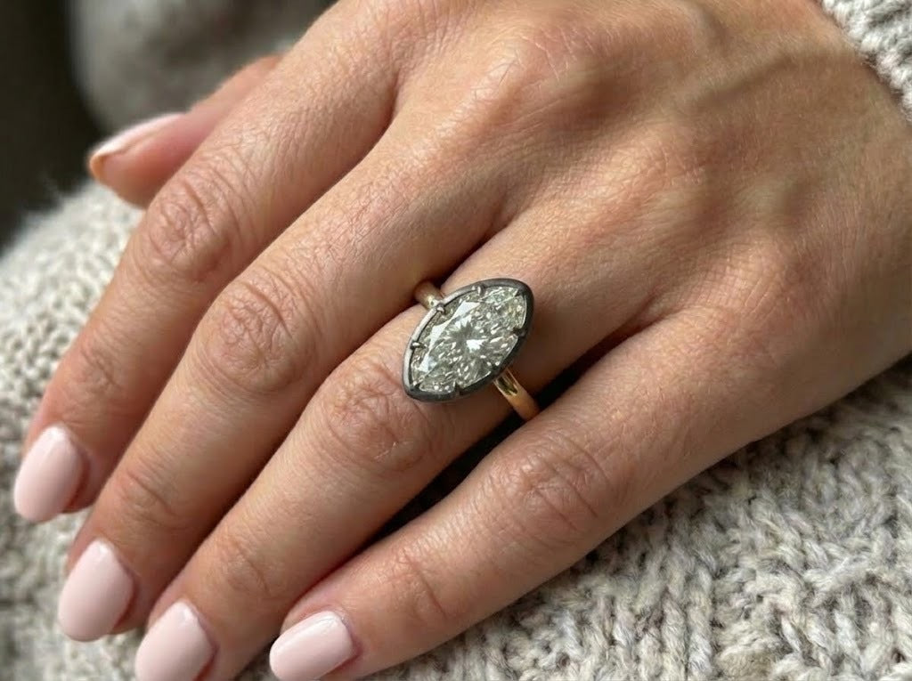 A hand with light pink manicured nails rests on knitted fabric, wearing the Collet Set Marquise Diamond Solitaire Ring in Black Rhodium and Yellow Gold, its marquise diamond beautifully catching the light.