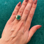 A hand with mauve manicured nails wears the Bezel Set Emerald Cut Emerald Engagement Ring in Yellow Gold and Platinum with Trapezoid Baguette Diamond Side Stones, set against a textured teal background.