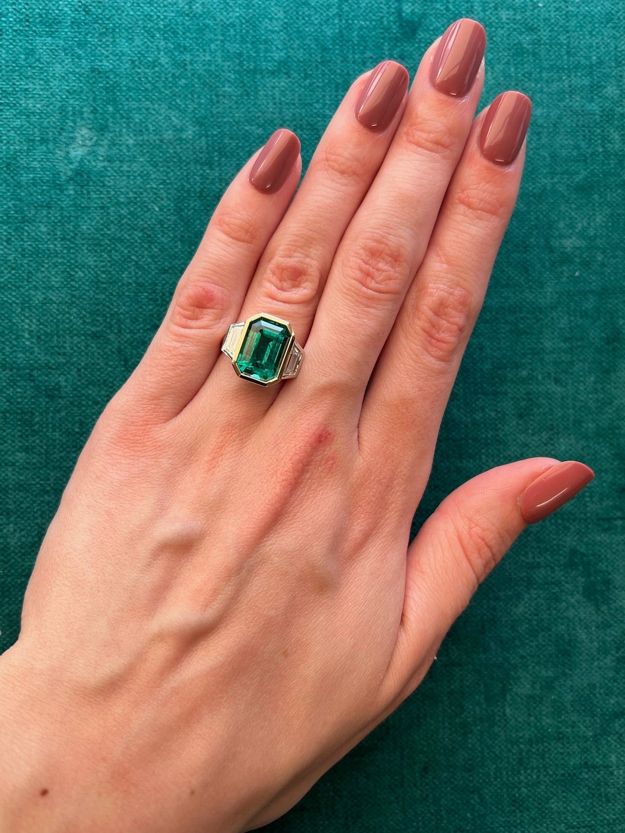 A hand with mauve manicured nails wears the Bezel Set Emerald Cut Emerald Engagement Ring in Yellow Gold and Platinum with Trapezoid Baguette Diamond Side Stones, set against a textured teal background.