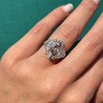 A hand with natural, nude-colored nails wears the Oval Pink Diamond Engagement Ring with Epaulette Diamond Side Stones in platinum and yellow gold against a green background.