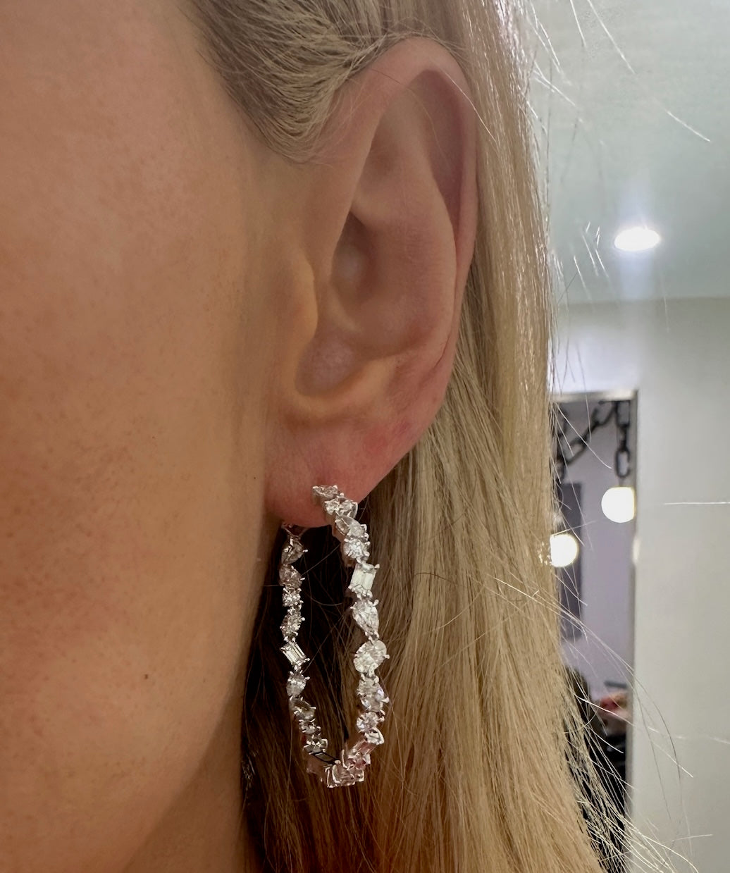 Close-up of a person with long blonde hair wearing the Mixed Shape Diamond Hoops, featuring dazzling diamonds in various shapes. The indoor background has blurred lights and a mirror.