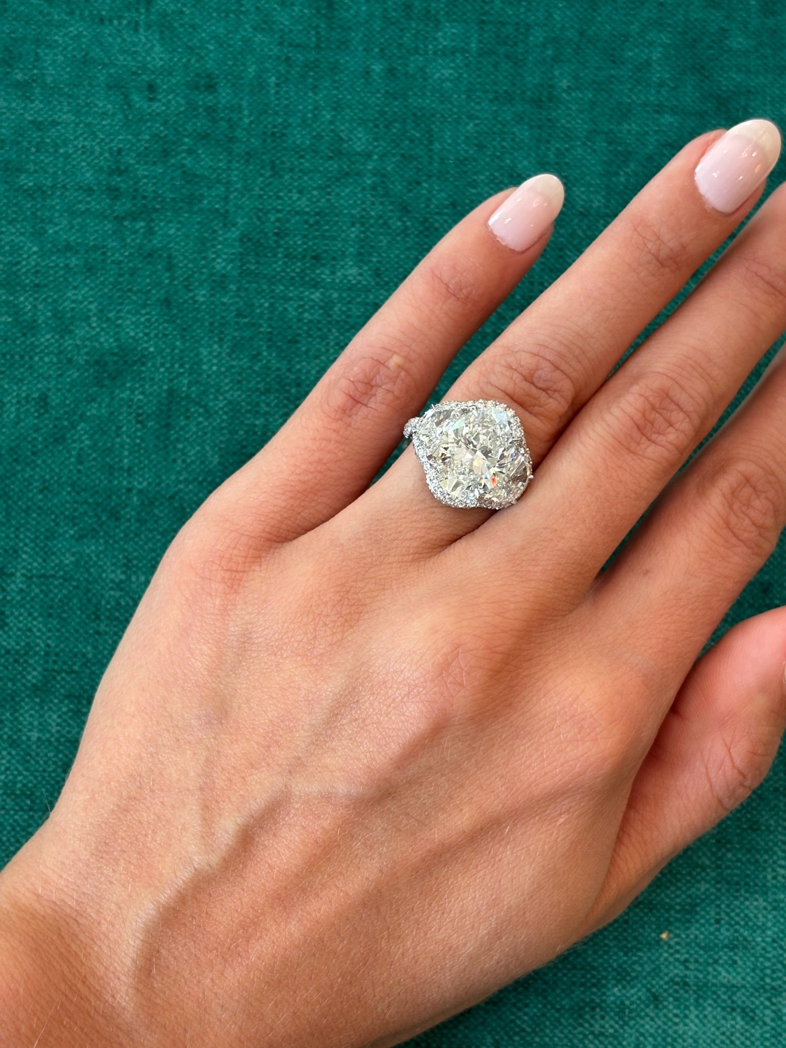 A hand with neatly manicured nails is shown against a green textured background, wearing the Three Stone Halo Oval Diamond Ring on the ring finger.