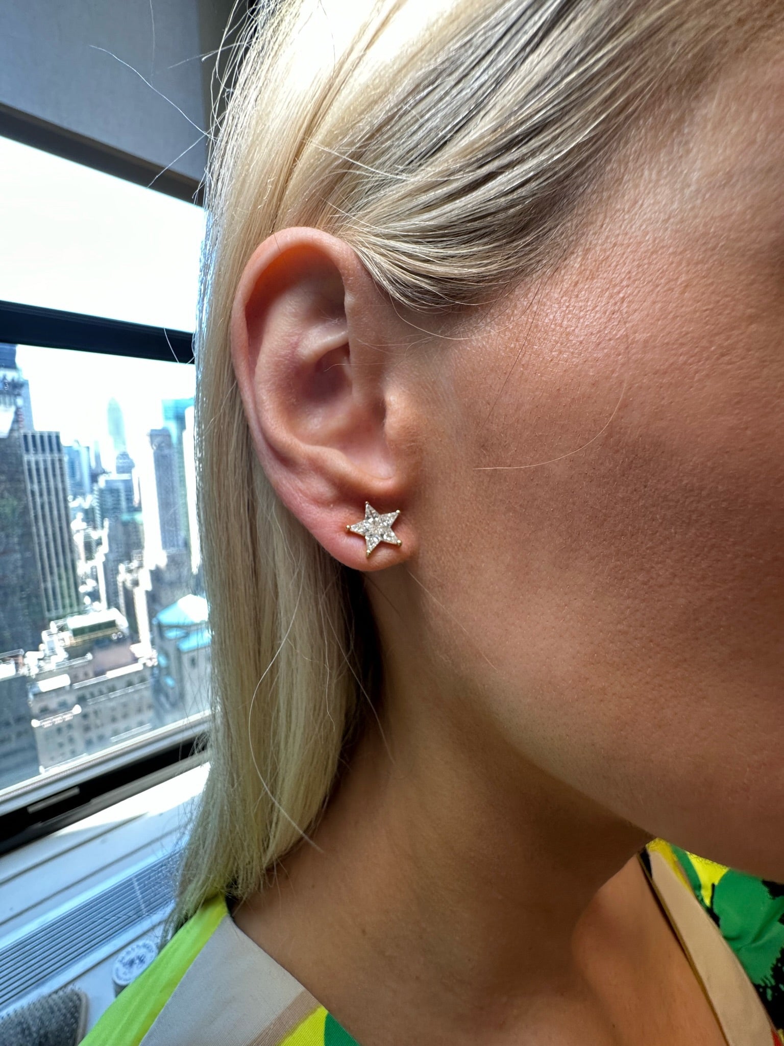 A close-up shows a person wearing Diamond Star Studs in Yellow Gold with sparkling gems, set against a cityscape visible through a window in bright daylight.