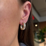 Close-up of a person wearing East-West Half-Bezel Set Emerald Cut Diamond Hoops in yellow gold, featuring clear emerald-cut stones. Part of the ear, cheek, and brown hair are visible against a blurred green and indoor-lit background.
