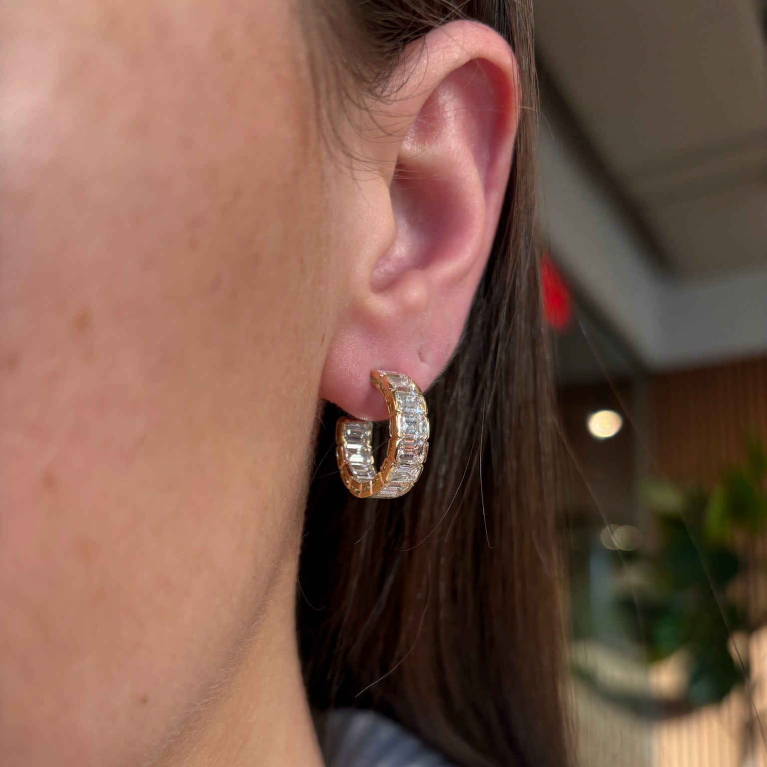 Close-up of a person wearing East-West Half-Bezel Set Emerald Cut Diamond Hoops in yellow gold, featuring clear emerald-cut stones. Part of the ear, cheek, and brown hair are visible against a blurred green and indoor-lit background.