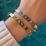 A hand wears two Burnish Set Mixed Shape Diamond Bangles—one 18K gold, one silver—with sparkling mixed shape diamonds. The person is dressed in a cream knit sweater against a teal fabric background.