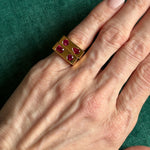 A hand with light skin wears the Mixed Shape Ruby Bar Ring in Yellow Gold, featuring six oval red gemstones, displayed against a green fabric background.