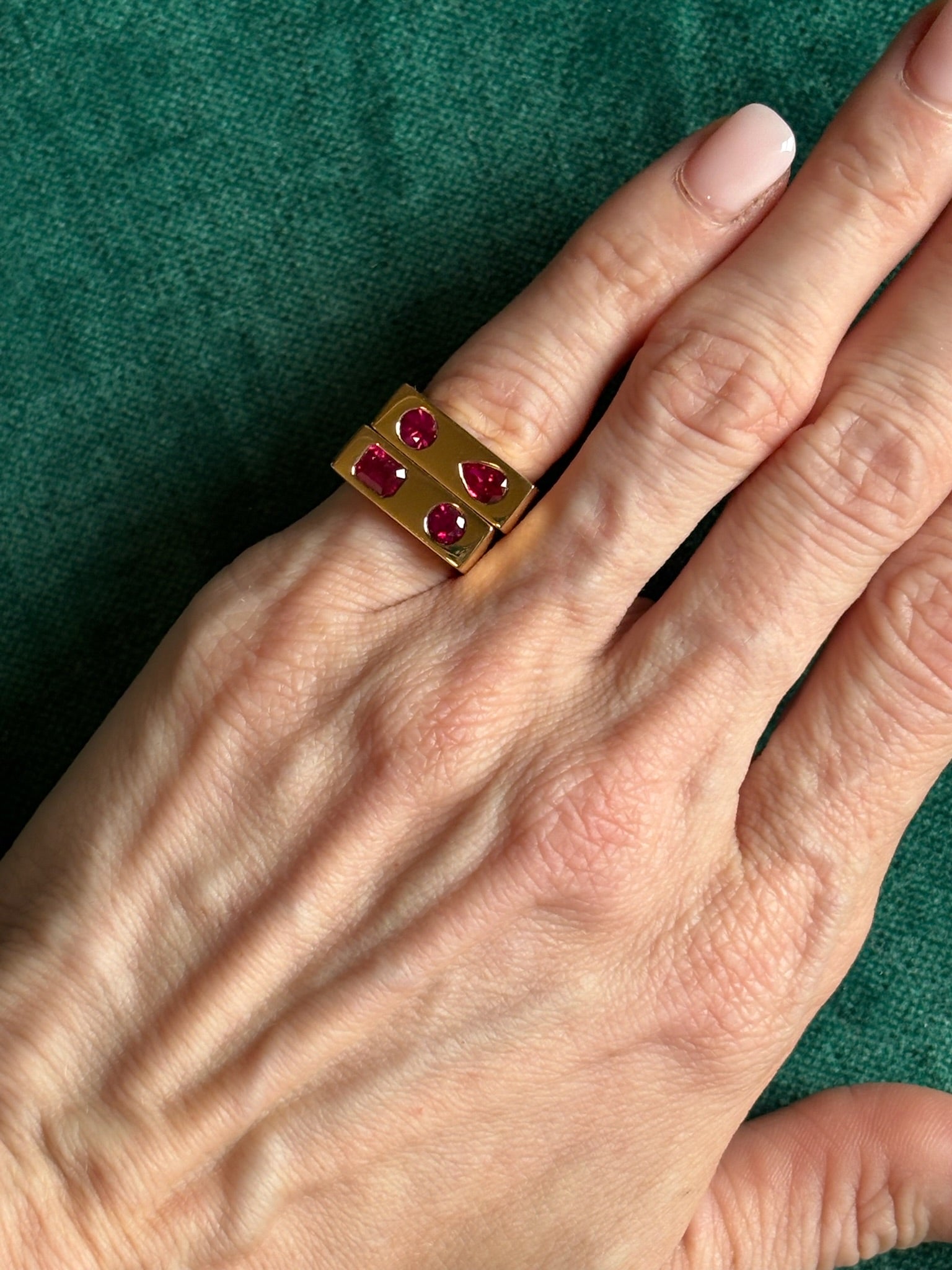 A hand with a light skin tone wears the Bar Ring with Round and Pear Cut Rubies in Yellow Gold against a green fabric background.