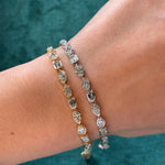A close-up of a wrist wearing the East-West Bezel Set Mixed Shape Diamond Tennis Bracelet in yellow gold against green fabric, featuring stunning oval and rectangular diamonds in a modern tennis bracelet design.