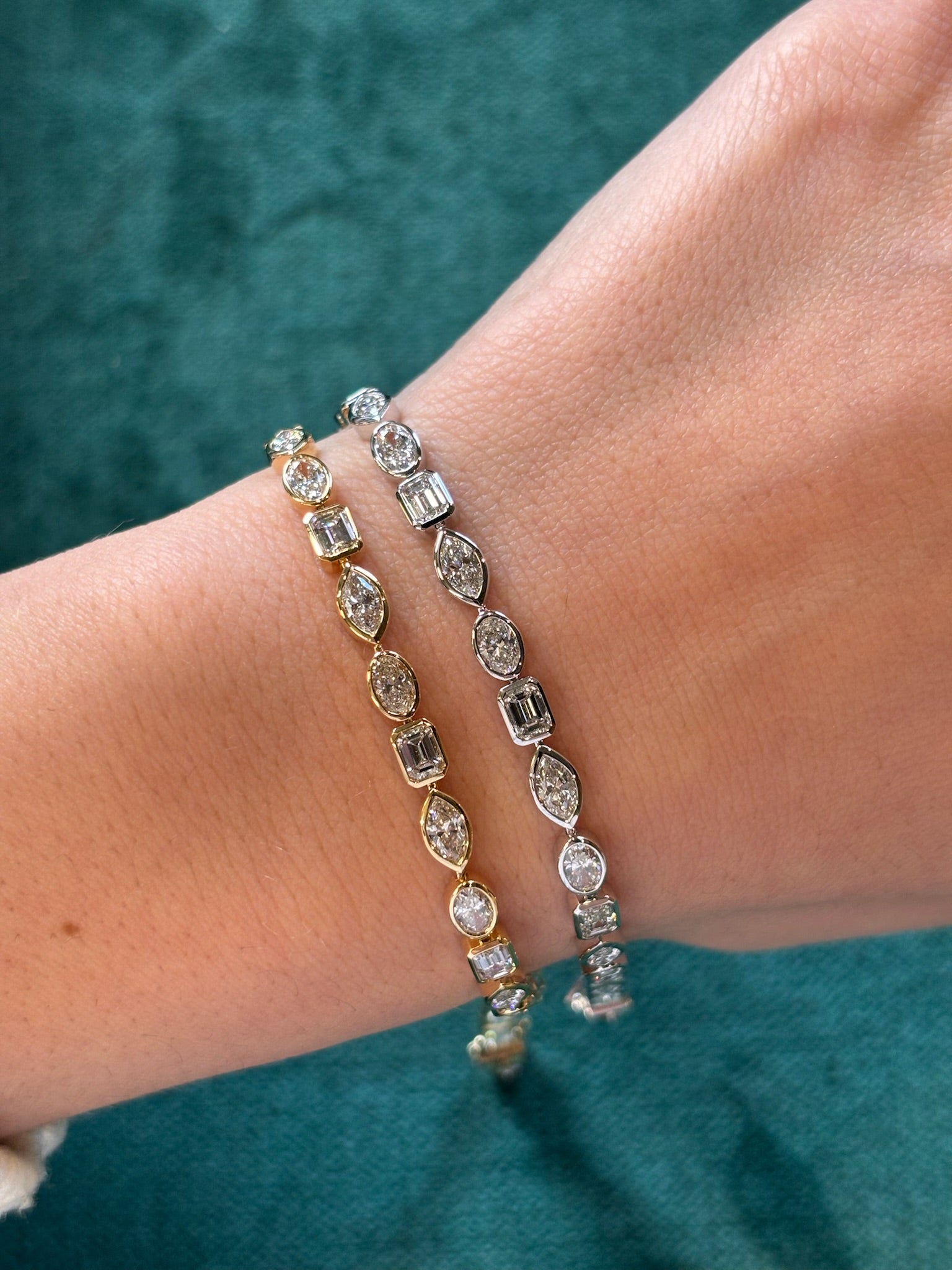 A close-up of a wrist wearing the East-West Bezel Set Mixed Shape Diamond Tennis Bracelet in yellow gold against green fabric, featuring stunning oval and rectangular diamonds in a modern tennis bracelet design.