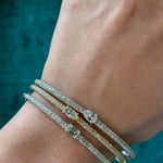 A hand wears three sparkling bangles—including the East-West Set Oval Shape Diamond Pavé Bangle—each with unique diamond accents, set against a teal textured background.