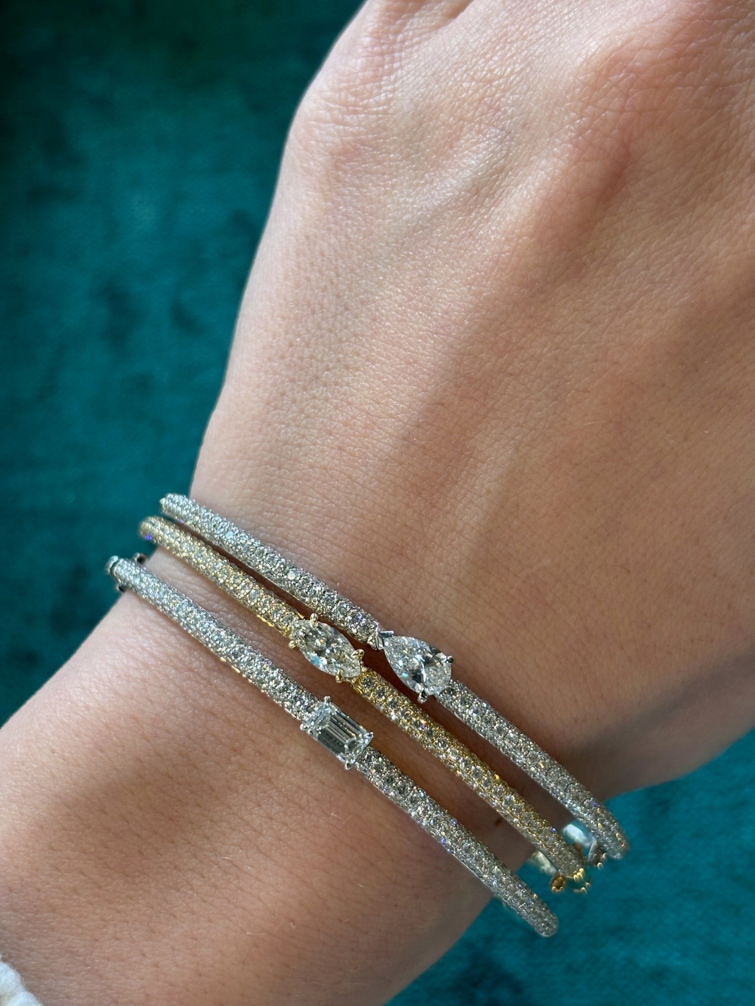 A hand wears three sparkling bangles—including the East-West Set Oval Shape Diamond Pavé Bangle—each with unique diamond accents, set against a teal textured background.