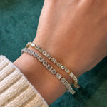 A hand in a cream-colored knit sweater displays the Alternating East-West Set Round and Emerald Cut Diamond Tennis Bracelet in White Gold against a green background.