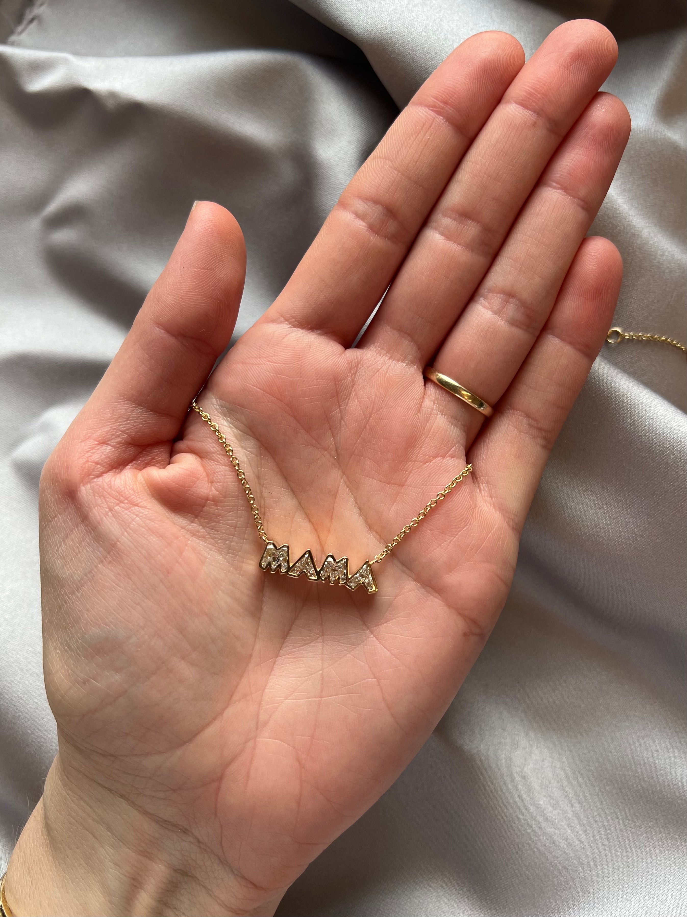 A hand with a gold ring holds the Bezel Set "Mama" Diamond Pendant Chain Necklace in Yellow Gold, featuring capital diamond letters, displayed against a light satin fabric background.