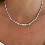 A close-up of a person wearing a white top and the Medium Graduated Round Diamond Tennis Necklace—an elegant necklace featuring round diamonds in a single row around their neck, radiating timeless sophistication.