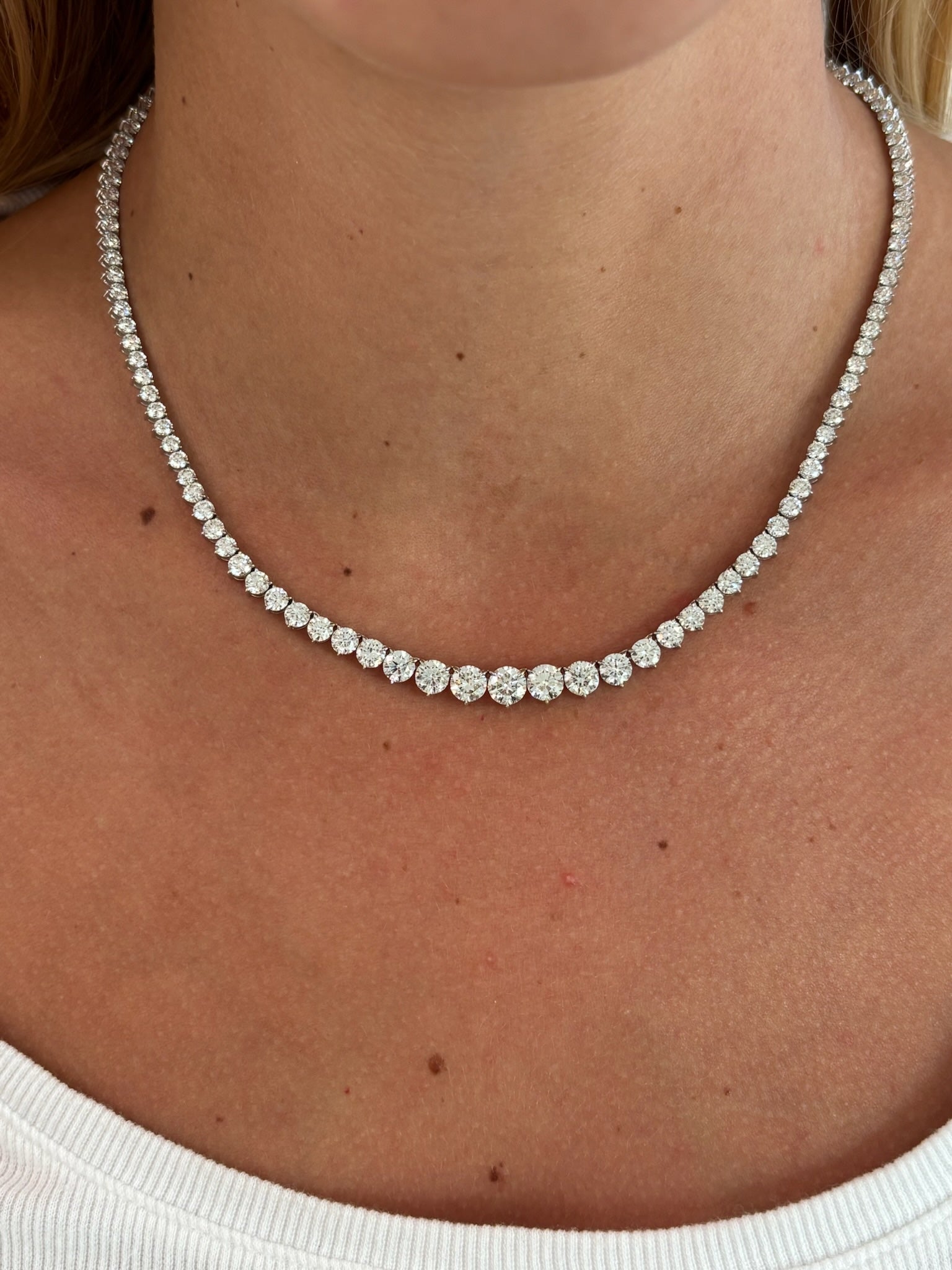 A close-up of a person wearing a white top and the Medium Graduated Round Diamond Tennis Necklace—an elegant necklace featuring round diamonds in a single row around their neck, radiating timeless sophistication.