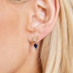 Close-up of a woman's ear wearing Double Kite Drops in Yellow Gold with Diamonds and Sapphires: one small gold-framed diamond stud and a larger drop earring with kite-shaped sapphires set in yellow gold.