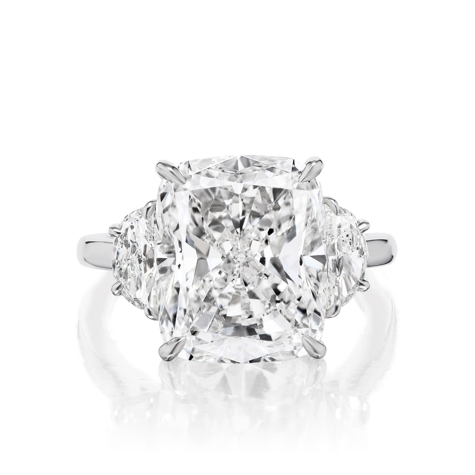 The Cushion Cut Diamond Engagement Ring with Half Moon Side Stones features a large cushion-cut center diamond flanked by two half moon diamonds, all set in a gleaming platinum band and displayed on a reflective white surface.