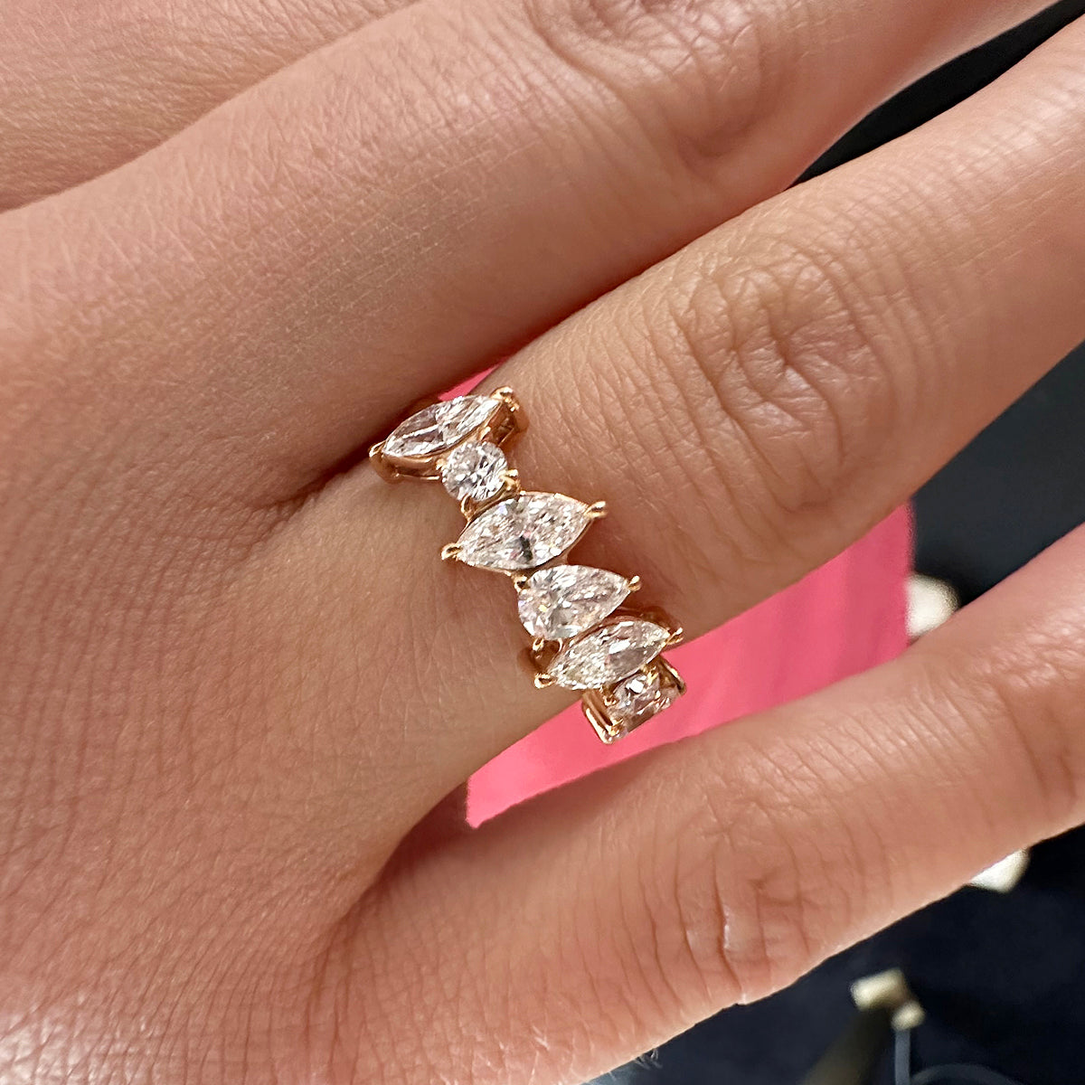 A close-up of a hand wearing the Diagonally Set Mixed Shape Diamond Half Eternity Band in 18K rose gold, featuring five marquise-cut diamonds, shown against a backdrop with a touch of pink fabric.