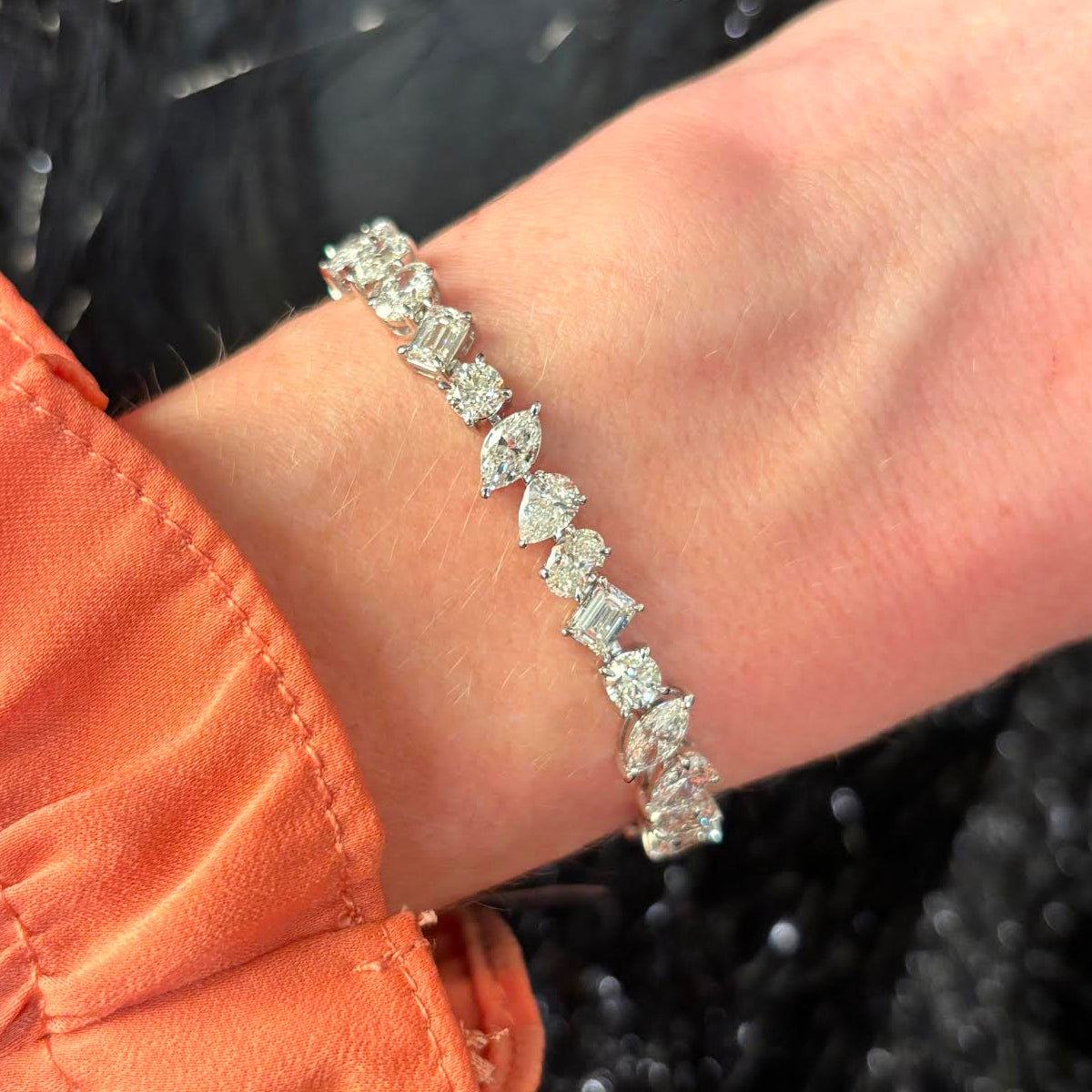 A close-up shows a wrist wearing the Mixed Shape Diamond Tennis Bracelet in White Gold, its sparkling diamonds catching the light against an orange long-sleeve top and a dark, textured background that highlights its brilliance.