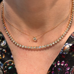 A close-up of a person's neck wearing two necklaces: a delicate 18K yellow gold chain with a diamond pendant and the Medium Hexagon Bezel Set Diamond Tennis Necklace in Yellow Gold, over a colorful floral-patterned top.
