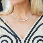 A woman in a cream dress with black swirls is shown from the shoulders up, wearing the Mixed Shape Diamond Tennis Necklace in White Gold around her neck.