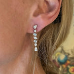 A close-up shows a woman's ear wearing Tennis Drop Earrings in White Gold with Mixed Shape Diamonds. Her blonde hair is partially visible, set against a colorful, blurred wall mural background.