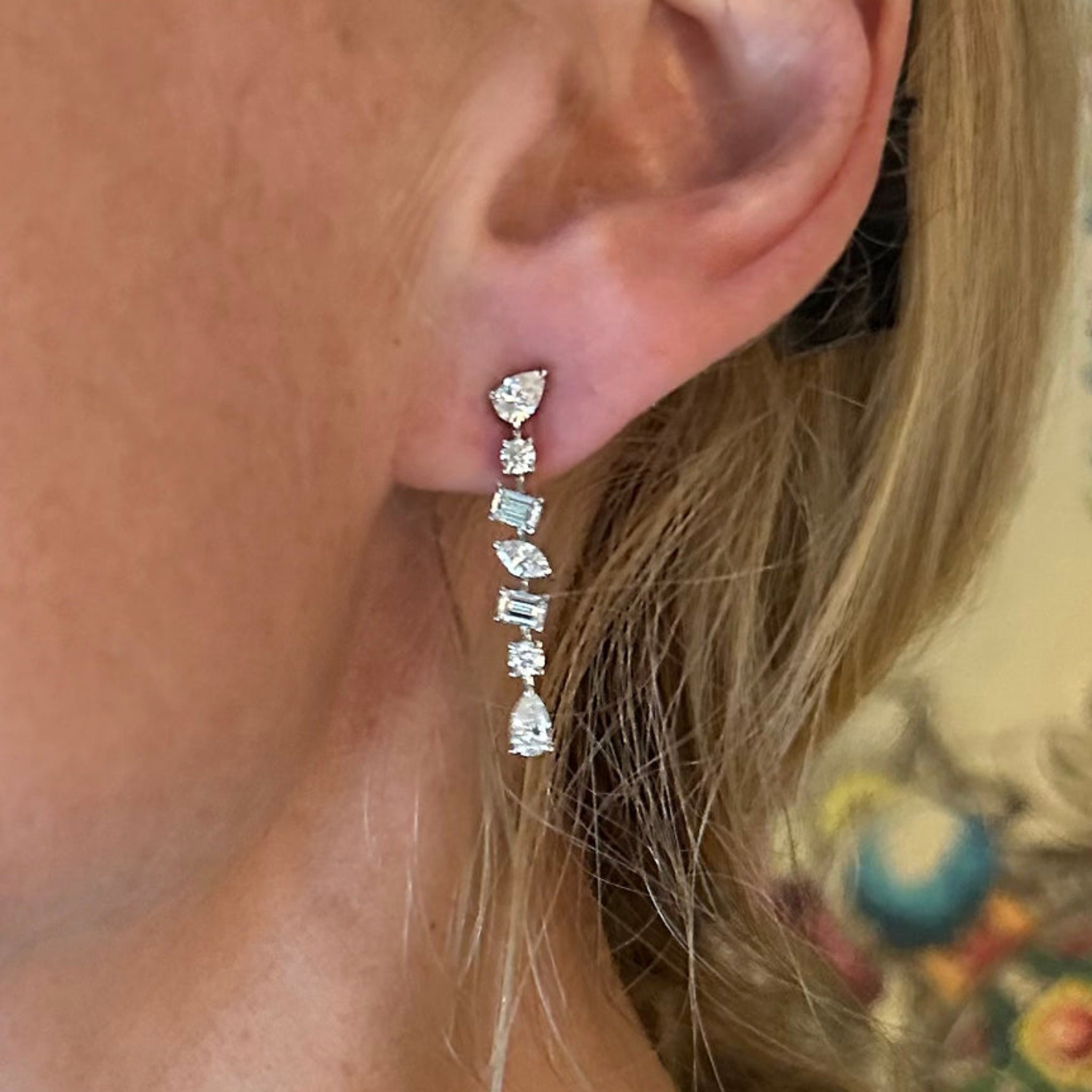 A close-up shows a woman's ear wearing Tennis Drop Earrings in White Gold with Mixed Shape Diamonds. Her blonde hair is partially visible, set against a colorful, blurred wall mural background.