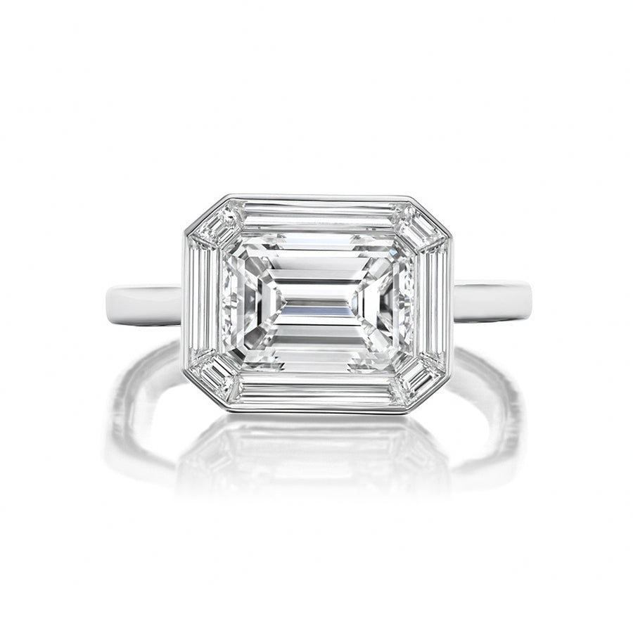 The East-West Mosaic Ring features baguette and emerald-cut diamonds set in white gold or platinum, highlighted by a large rectangular emerald-cut center stone, shown from the front on a white background.