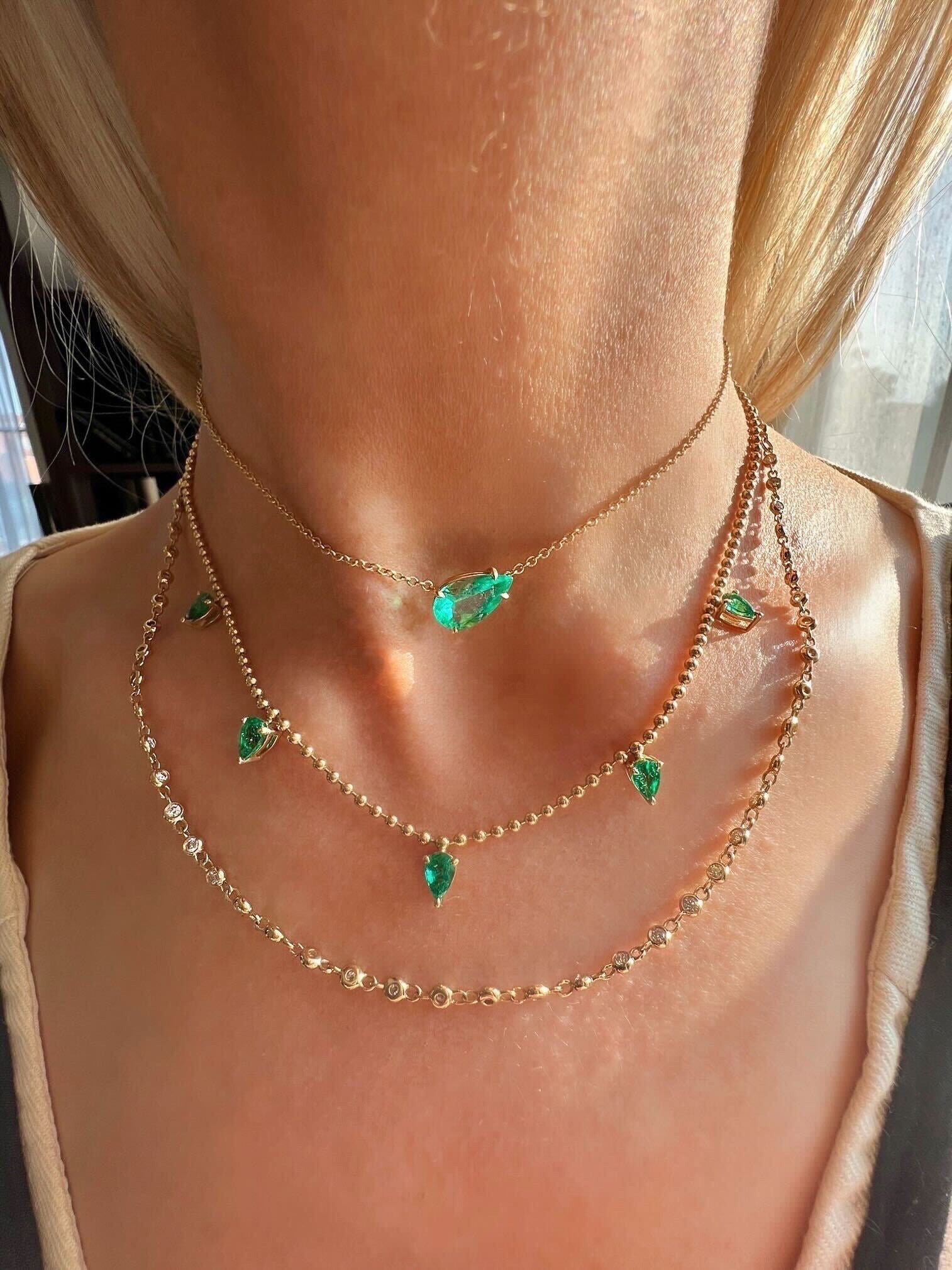 A close-up of a fair-skinned person with light blonde hair wearing three layered gold necklaces, featuring the Golden Garland Necklace in yellow gold with round brilliant diamonds and green gemstone pendants reflecting sunlight.