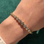 The Graduated Hexagon Bezel Set Diamond Tennis Bracelet, crafted in 18K gold, showcases clear round brilliant diamonds nestled in hexagonal bezel settings and is elegantly worn on a person’s wrist against a green fabric background.