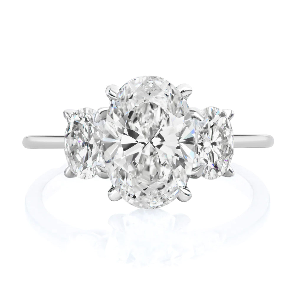 The Oval Diamond Engagement Ring with Oval Side Stones features a platinum band with a large oval-cut diamond center and two smaller ovals on each side—an elegant, timeless choice for a custom design.