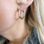 A close-up shows a person wearing Chrysler Hoops with Baguette Diamonds—gold hexagonal earrings featuring baguette diamonds. Their blonde hair and dark-colored top are also visible.