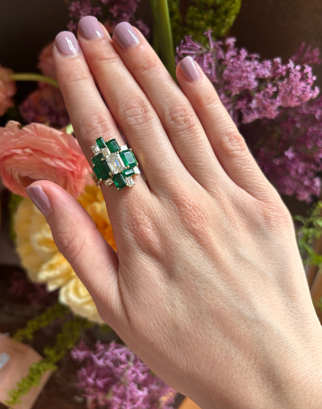 A hand with light pink nails showcases the Bespoke Geometric Emerald Cut Emerald and Diamond Ring Jacket in Yellow Gold with Pavé Band, set against assorted pink and yellow flowers.