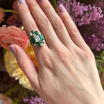 A hand with light purple nail polish wears a Geometric Emerald Cut Emerald and Diamond Ring Jacket in Yellow Gold with Pavé Band. A large ring with green and white gemstones is paired, set against a backdrop of pink, purple, and yellow flowers.