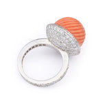 The Cartier Carved Coral and Diamond Ring, circa 1950s, features a large, round, ribbed orange coral cabochon set atop a silver band encrusted with pavé diamonds.