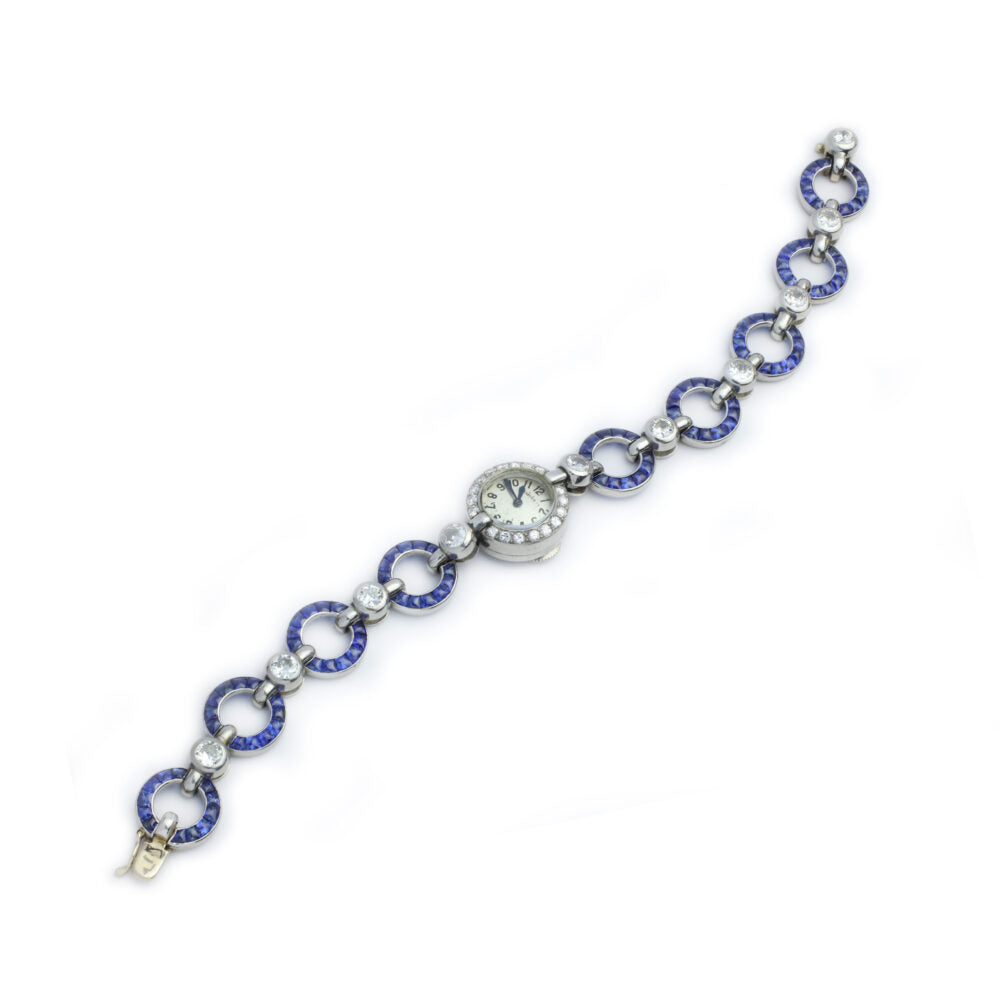 The Cartier Art Deco Sapphire and Diamond Watch features a circular silver face set in a bracelet of alternating silver and sapphire-studded rings, linked by small diamond accents.