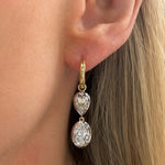 Close-up of a person's ear wearing the Collet Set Oval Shape and Pear Shape Earring in White Gold and Yellow Gold, with two dangling clear gemstones—one teardrop and one oval. Blonde hair is visible on the right side.