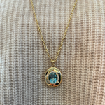 The Pavé Oval Cut Aquamarine Collet Pendant in Yellow Gold is worn over a cream-colored knitted sweater.